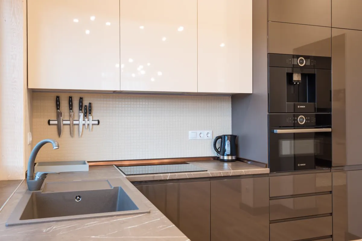 Glossy cream and taupe kitchen cabinets with a mosaic tile backsplash and built-in ovens